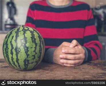 A young man is sitting at a table in a kitchen with a fresh watermelon next to him