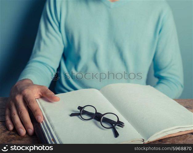 A young man is sitting at a table with a big notepad and a pair of glasses in front of him