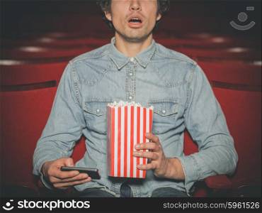 A young man is sitting in a cinema and is watching a film while holding his smartphone and a bucket of popcorn