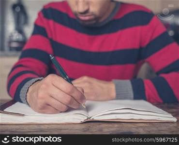 A young man is sitting in a kitchen and is writing in a big notebook