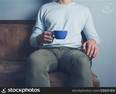 A young man is sitting on a sofa and is drinking coffee from a blue cup