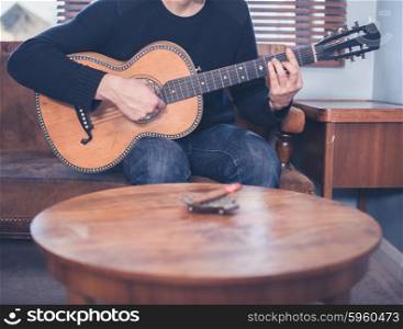 A young man is sitting on a sofa at home in his living room and is playing guitar, there is a coffee table with an ashtray and a cigar on it in the foreground