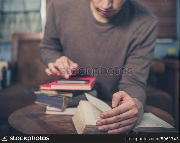 A young man is sitting on a sofa surrounded by books and is using his smartphone