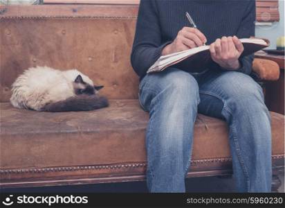 A young man is sitting on a sofa with a cat and is writing in a notebook