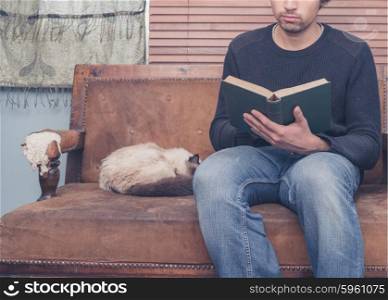 A young man is sitting on a sofa with a cat and is reading a big book