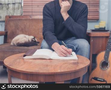 A young man is sitting with his cat and guitar at home on a sofa and is writing songs in a notebook