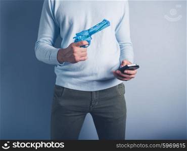 A young man is standing against a blue background with a water pistol and a smart phone