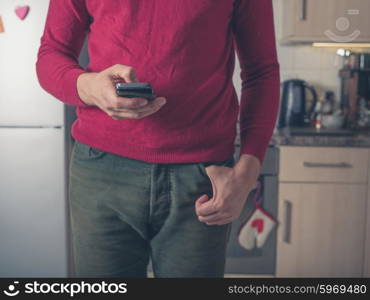 A young man is standing in a kitchen and is using a smart phone