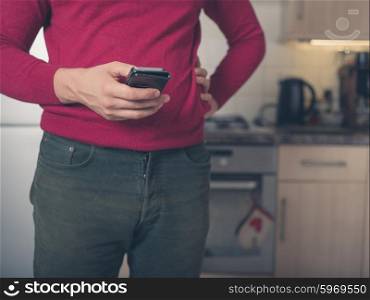 A young man is standing in a kitchen and is using a smart phone