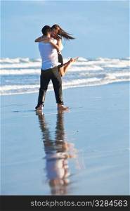 A young man lifting and embracing his girlfriend barefoot on a beach with a bright blue sky