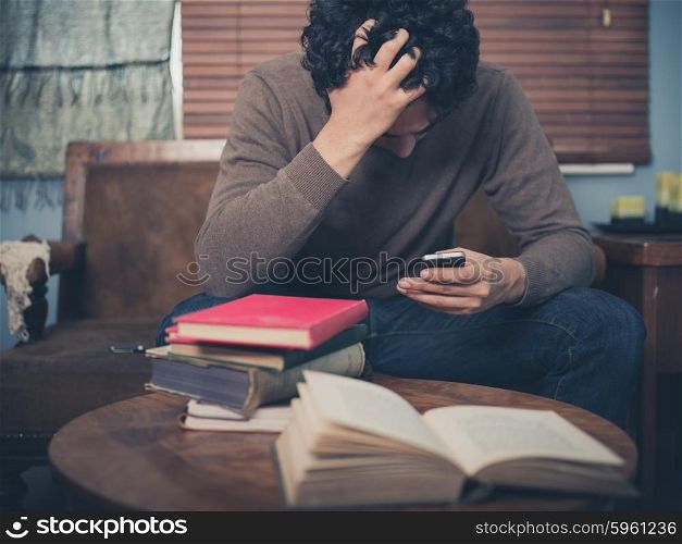 A young man surrounded by books is sitting on a sofa and is looking upset as he is using his smartphone