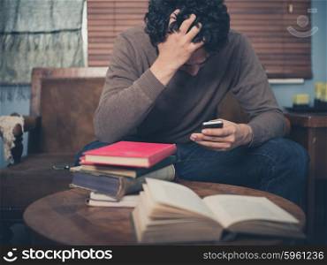 A young man surrounded by books is sitting on a sofa and is looking upset as he is using his smartphone