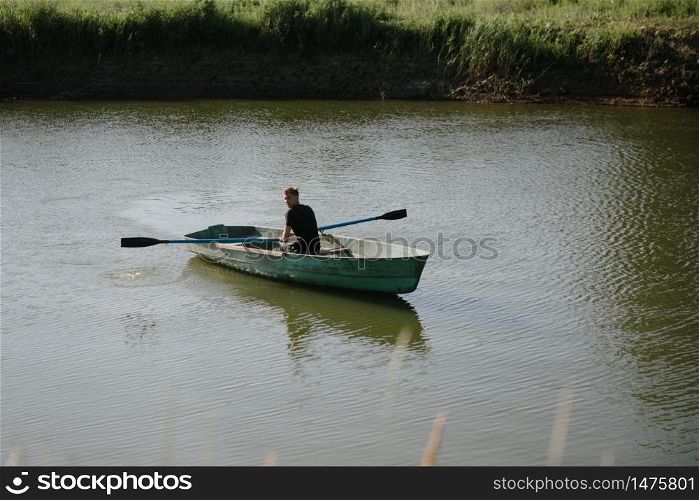 A young man swims in a small wooden boat with oars on the river. Young man floats on a wooden boat with oars