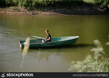 A young man swims in a small wooden boat with oars on the river. Young man floats on a wooden boat with oars