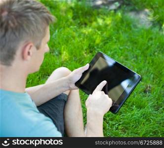 A young man uses tablet computer relaxing in green grass
