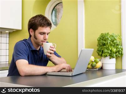 A young man uses the computer in the kitchen while enjoying a warm drink.