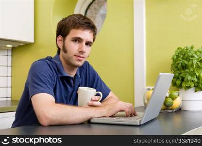A young man uses the computer in the kitchen while enjoying a warm drink