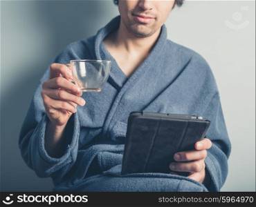 A young man wearing a bathrobe is holding an empty coffee cup and is using a tablet