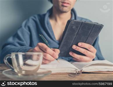 A young man wearing a bathrobe is sitting at a table and is using a tablet while taking notes in a book