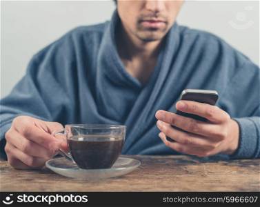 A young man wearing a blue bathrobe is sitting at a table and is using a smart phone while having a cup of coffee in the morning