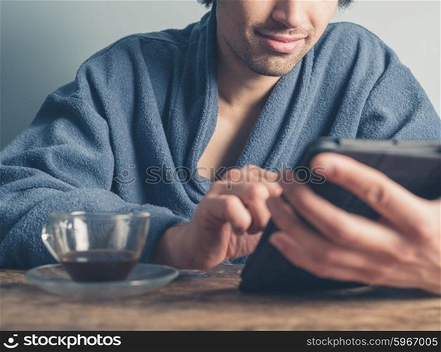 A young man wearing a blue bathrobe is sitting at a table and is using a tablet while having a cup of coffee