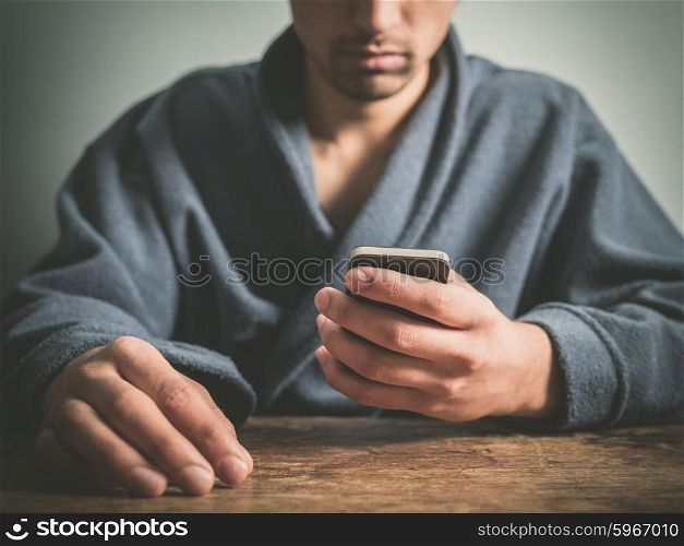 A young man wearing a blue bathrobe is sitting at a table and is using a smart phone