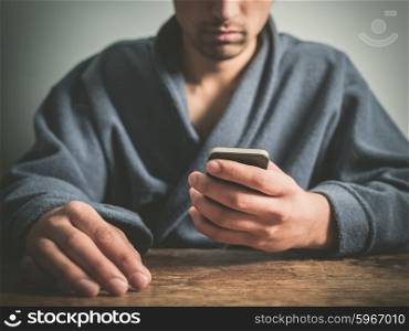 A young man wearing a blue bathrobe is sitting at a table and is using a smart phone