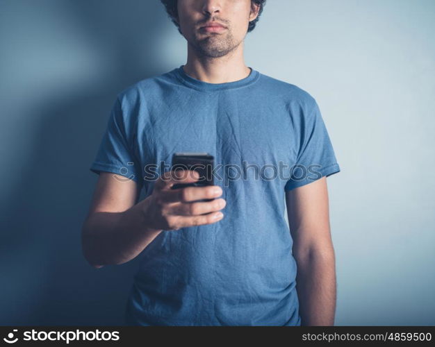 A young man wearing a blue t-shirt is holding a smartphone by a blue wall