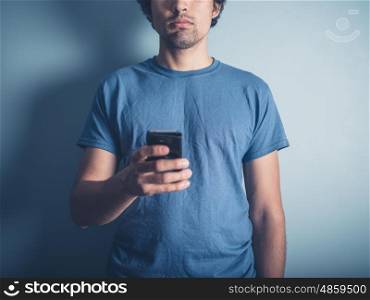 A young man wearing a blue t-shirt is holding a smartphone by a blue wall