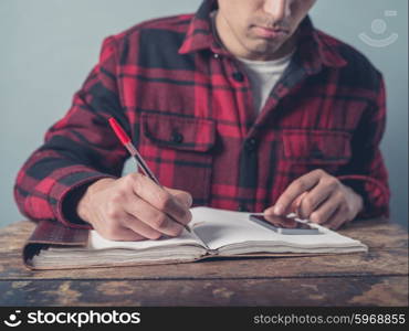 A young man wearing a checkered jacket is using a smartphone and is taking notes in a notebook at a desk