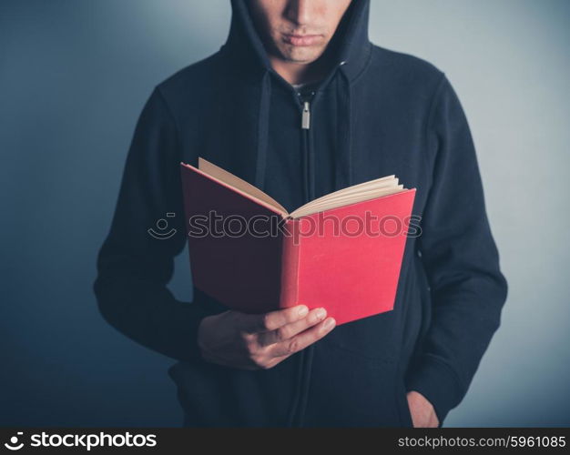 A young man wearing a hooded top is standing around and is reading a red book