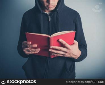 A young man wearing a hooded top is standing around and is reading a red book