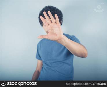 A young man wearing blue is raising his hand to cover and hide his face
