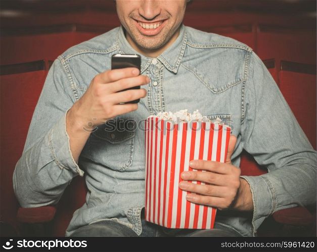 A young man with a bucket of popcorn is sitting in a cinema and is using his smartphone