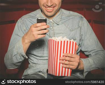 A young man with a bucket of popcorn is sitting in a cinema and is using his smartphone