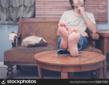 A young man with bare feet is resting on a sofa at home with a cat sleeping next to him