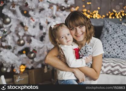 A young mother gently hugs the little girl before Christmas.. Family hug of a mother and daughter in front of a Christmas tree 7121.