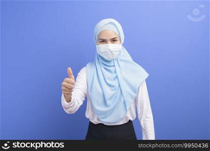 A young muslim woman with hijab wearing a surgical mask over blue background studio.. Young muslim woman with hijab wearing a surgical mask over blue background studio.