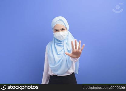 A young muslim woman with hijab wearing a surgical mask over blue background studio.. Young muslim woman with hijab wearing a surgical mask over blue background studio.