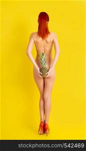 A young naked girl wearing only high-heeled shoes stands with her back to the viewer closing her ass with pineapple fruit on a bright yellow background. girl With Pineapple