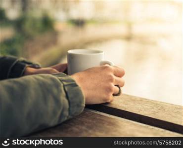 A young persons hands holding a mug with a hot beverage by a pond in a park at sunset in the winter