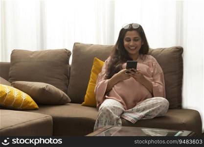 A YOUNG PREGNANT WOMAN SITTING ON A SOFA AND HAPPILY USING MOBILE PHONE