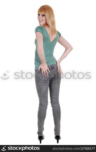 A young pretty and tall woman in jeans and green sweater standing from the back in the studio, holding her butt, over white background.