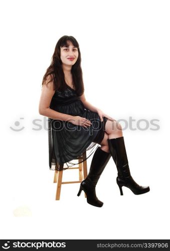 A young pretty woman sitting in a black blue dress and boots ona chair and smiling into the camera for white background.