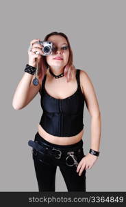 A young punk woman standing in black leather pants and a black corset witha camera in her hand for gray background.