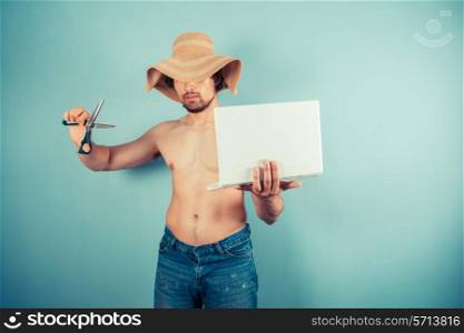 A young shirtless man wearing a beach hat is about to cut his laptop with a pair of scissors