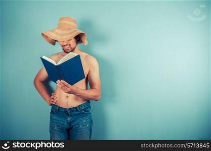 A young shirtless man wearing a large beach hat is reading a book