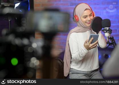 A young smiling muslim female singer wearing headphones with a microphone while recording song in a music studio with colorful lights.. Young smiling muslim female singer wearing headphones with a microphone while recording song in a music studio with colorful lights.