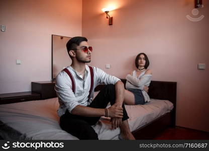 a young stylish guy in glasses and a young girl together sitting on the bed. the guy closer to the girl further. selective focus.