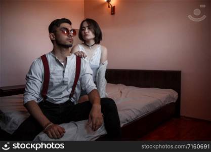 a young stylish guy in glasses and a young girl together sitting on the bed. the guy closer to the girl further. selective focus.